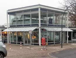 Das Foto zeigt ein modernes, transparent gestaltetes Geb�ude in Hamburg-Rissen. Es handelt sich um einen denkmalgesch�tzten Glaspavillon mit zwei Etagen. Die Struktur besteht aus einem wei�en Stahlrahmen und gro�fl�chigen Glasfronten, die den Blick ins Innere und auf eine Treppe im Erdgeschoss freigeben. Ein schmales Glasvordach uml�uft das Erdgeschoss. Das Geb�ude steht an einer gepflasterten Stra�enecke; im Vordergrund sind rot-wei�e Absperrpfosten und ein Stra�enschild mit der Aufschrift �Wedeler Landstra�e� zu sehen.