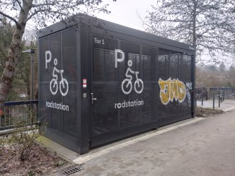 Schwarze, moderne Radstation (Fahrradabstellanlage) der S-Bahn-Station Rissen. Das eingeschossige, containerartige Geb�ude mit gro�fl�chigen Gitterverkleidungen und Glast�ren steht ebenerdig direkt neben einem gepflasterten Gehweg. Der Zugang erfolgt �ber eine flache, stufenlose Eingangst�r mit T�rdr�cker in normaler H�he. Der Boden vor und im Eingangsbereich ist eben und ohne Schwellen. Rechts neben dem Eingang befindet sich eine Bedientafel in typischer H�he. Die Station ist beleuchtet und verf�gt �ber einen breiten Zugangsbereich. Im Hintergrund f�hrt ein asphaltierter Weg weiter, der ebenfalls stufenlos und gut befahrbar ist.