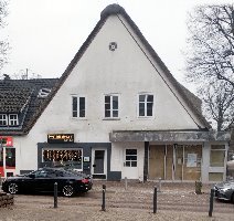 Zweigeschossiges Geb�ude mit wei�er Fassade und steilem Giebeldach an einer Stra�e. Im Erdgeschoss befinden sich mehrere Eing�nge und Fenster, links ein Laden mit beleuchteter Dekoration. Vor dem Geb�ude sind parkende Autos und einige Poller zu sehen, rechts ein Baum ohne Bl�tter.