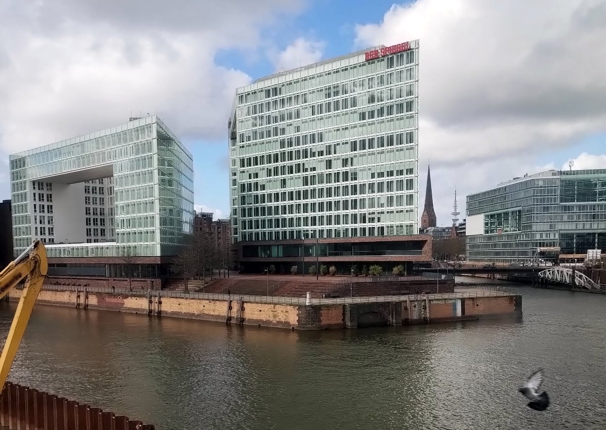 Moderne Glas- und Stahlhochhäuser an einem Hafenbecken in einer Großstadt unter blauem, leicht bewölktem Himmel. Links ein markantes, U-förmiges Glasgebäude, rechts ein hoher rechteckiger Glas-Turm. Im Vordergrund ein Kanal mit Steinpromenade, ein gelber Baggerarm links sowie eine fliegende Taube unten rechts. Im Hintergrund Kirchturm und weitere moderne Gebäude.